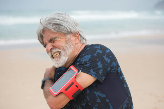 Free Elderly man grimacing from shoulder pain during seaside walk, showcasing health issue outdoors.